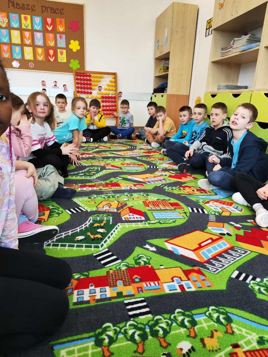Grupa uczniów klasy I siedząca na kolorowym dywanie o ruchu drogowym