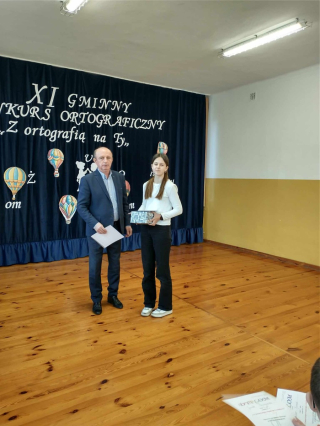 Dyrektor wręczający nagrodę uczniowi w konkursie na granatowym tle z napisem XI Gminny Konkurs Ortograficzny „Z ortografią na TY ”