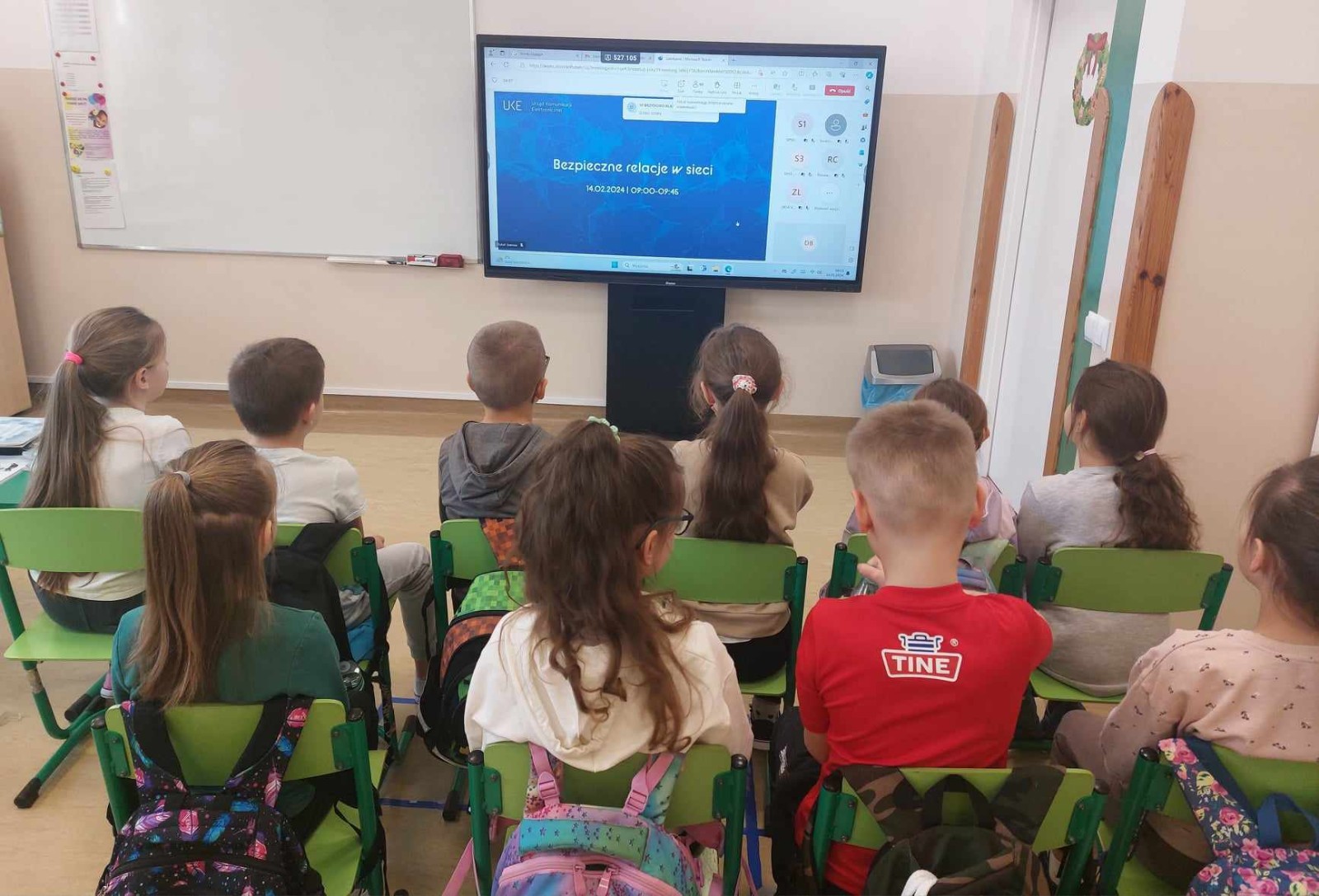 Grupa uczniów siedząca na krzesełkach i oglądająca na monitorze interaktywnym webinar Bezpieczne relacje w sieci