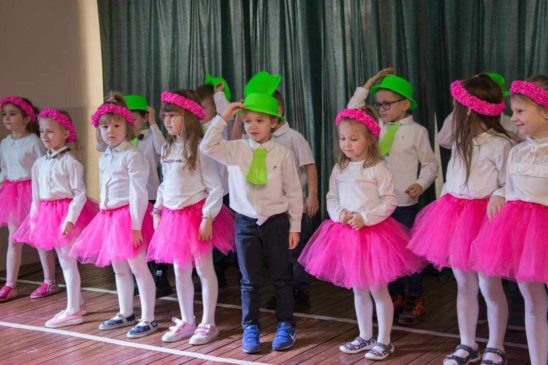 Grupa przedszkolaków w różowych spódniczkach i białych bluzkach na scenie
