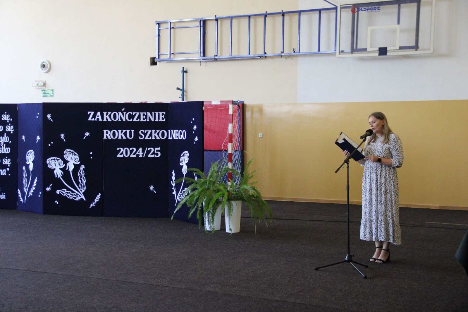 Zakończenie roku przedszkolnego - prowadząca na tle dekoracji na niebieskim tle przemawiająca do mikrofonu