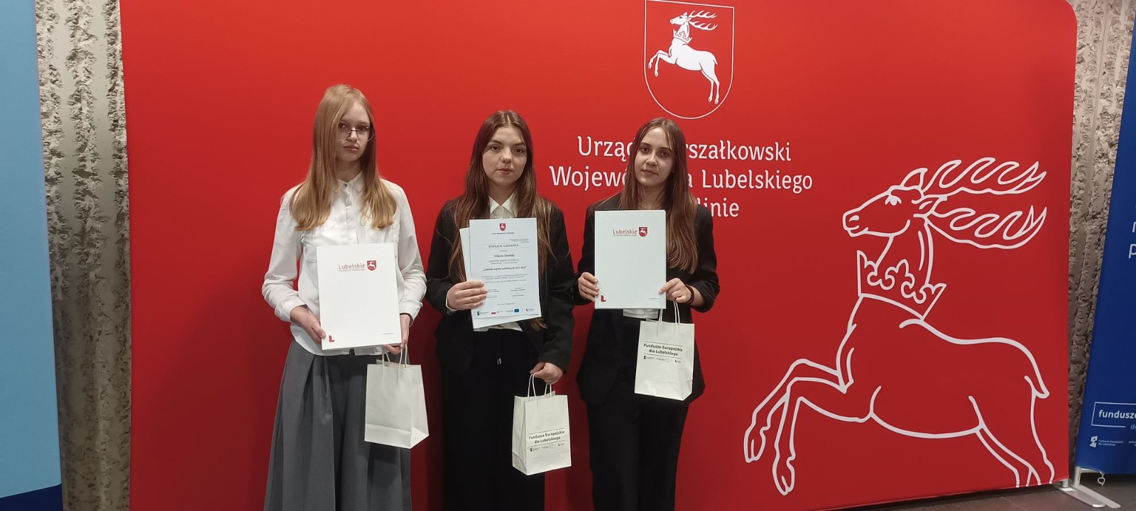 Trzy dziewczyny pozują na tle czerwonej ścianki z logo Województwa Lubelskiego (biały jeleń w koronie). Każda z nich trzyma białą teczkę z napisem „Lubelskie” oraz małą papierową torebkę upominkową. Dziewczyny są ubrane w stroje galowe.